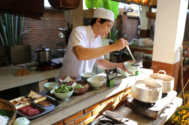 Đầu bếp mặc đồng phục trắng chuẩn bị món phở tươi tại bếp ngoài trời với rau thơm và nồi nước dùng ở Hội An.