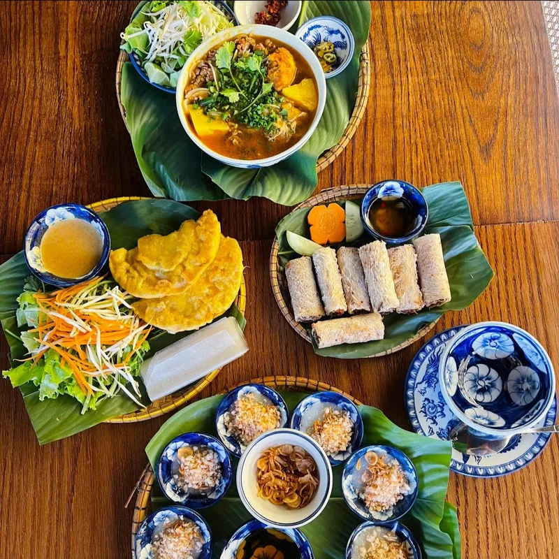 Bàn gỗ ở Huế bày bún bò Huế, bánh chiên, nem cuốn và nhiều món nhỏ trong bát hoa văn xanh.