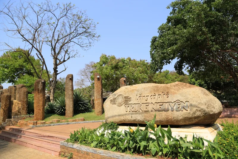 Tấm bia đá khắc chữ làng cà phê Trung Nguyên nổi bật giữa các cột đá và cây xanh ở Đắk Lắk.