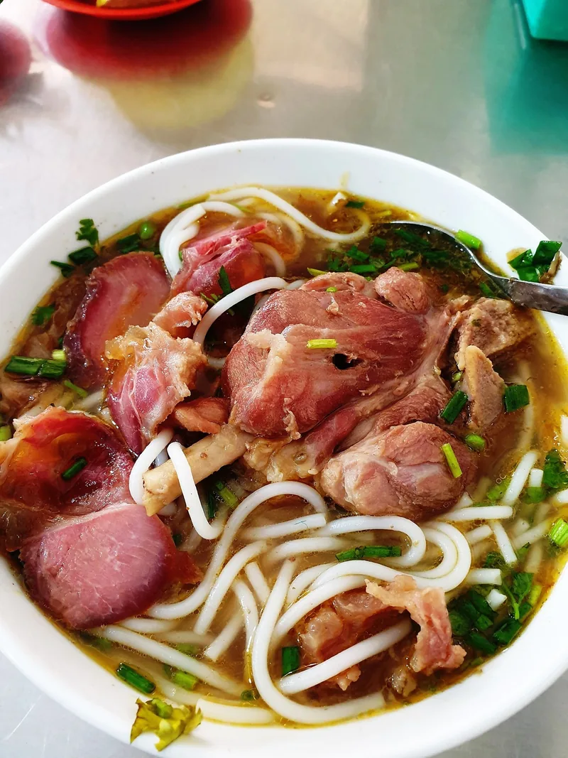 Một tô bún bò giò heo với sợi bún to, thịt giò heo và nước dùng điểm xuyết hành lá.