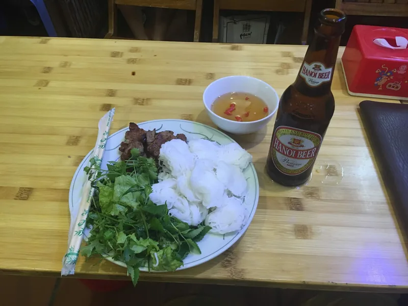 Đĩa chả nướng, rau thơm, bún cùng nước chấm và chai bia Hà Nội trên bàn gỗ.