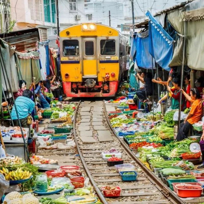 Đoàn tàu màu vàng chạy qua khu chợ đường tàu với các sạp rau củ tươi bày bán hai bên.