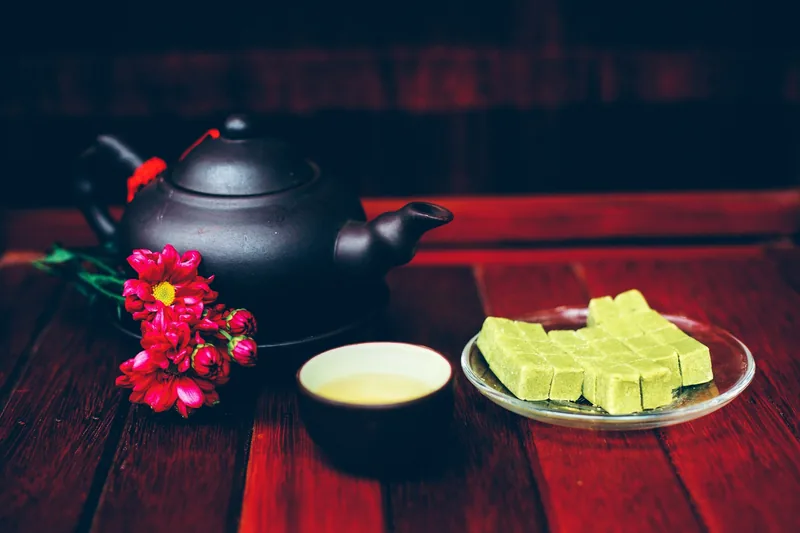 Ấm trà đất nung, chén trà, hoa đỏ và bánh đậu xanh bày trên bàn gỗ ở Việt Nam.