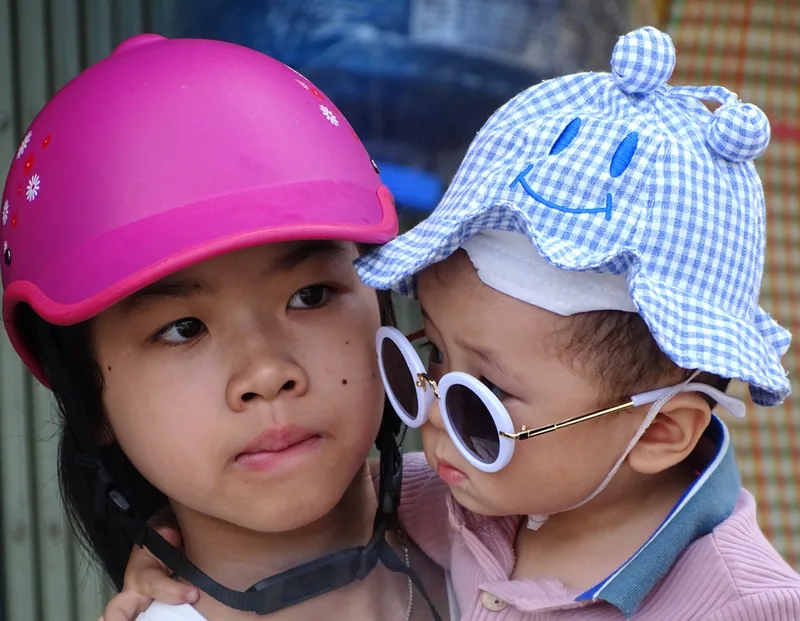 Một bé gái đội mũ bảo hiểm hồng bế em bé đeo kính râm và đội mũ hoa văn tại chợ phiên Bắc Hà gần Sa Pa.